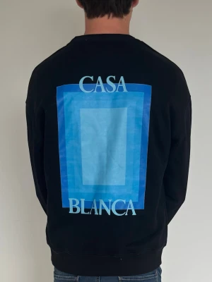 Svart sweatshirt från Casablanca - Cool svart sweatshirt från Casablanca med blått grafiskt tryck och texten 'Casa Blanca' på både bröstet och ryggen. Tröjan har rund halsringning och ribbade muddar vid ärmslut och nederkant. Perfekt för en avslappnad och trendig look.
