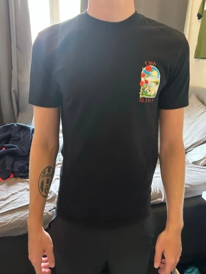 Svart Casa Blanca t-shirt med tryck - Snygg svart t-shirt från Casa Blanca med färgglatt tryck av blommor och landskap både fram och bak. T-shirten har rund hals och korta ärmar, perfekt för dig som vill sticka ut med en unik design.