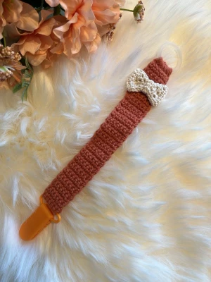 Virkad napphållare med rosett - Säljer en handgjord virkad napphållare i rostbrunt med en söt beige rosett. Hållaren har en plastklämma i orange och en genomskinlig ring för att fästa nappen. Perfekt accessoar för att hålla koll på nappen med stil.
