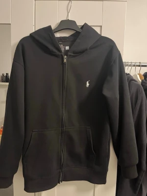Svart hoodie med dragkedja från Polo - Svart hoodie med dragkedja från Polo Ralph Lauren. Klassisk design med vit broderad logga på bröstet, två fickor fram och huva. Tillverkad i mjukt material som känns skönt mot huden. Perfekt för en avslappnad och stilren look.