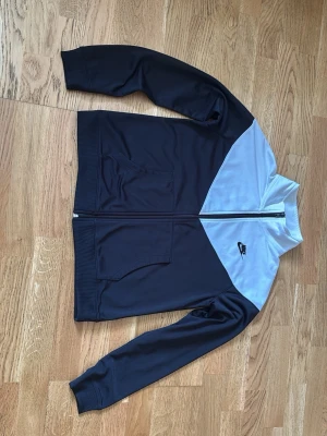 Nike svart och vit träningsjacka - Snygg träningsjacka från Nike i svart med vit panel upptill. Jackan har dragkedja framtill, två fickor och ribbade muddar vid ärmslut och nederkant. Perfekt för träning eller sportiga outfits.