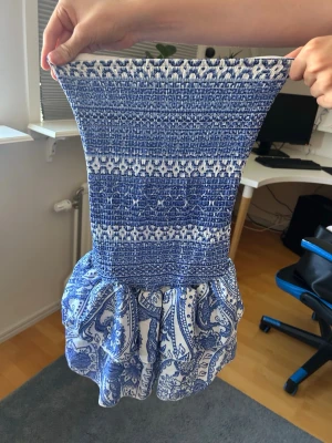 Blåvit kort klänning med volang - Säljer en blåvit kort klänning med smockad överdel och volang nertill. Klänningen har ett paisleyinspirerat mönster och är ärmlös, perfekt för varma dagar. Materialet känns lätt och luftigt, troligen bomull eller polyester. Supersnygg till sommaren!
