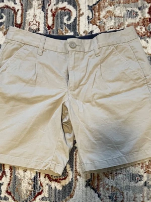 Beige shorts med fickor - Beige shorts med bälteshällor. De har två sidofickor fram och två passpoalfickor bak. Längd ytterben: 42 cm. Längd inre ben: 20 cm.