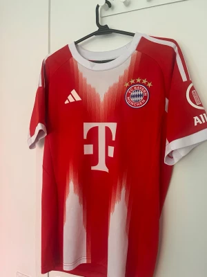 Röd Bayern München matchtröja Olise  - Säljer en röd och vit Bayern München matchtröja med klubbmärke och sponsortryck på bröstet. Tröjan har korta ärmar, rund hals och moderna detaljer i vitt längs sidorna och axlarna. Perfekt för fotbollsfans! Storlek S och ganska bra skick. Hör av er vid funderingar!