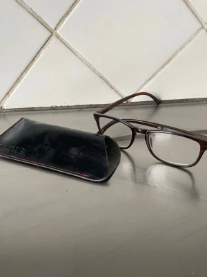 Foster Grant Bruna rektangulära glasögon - Snygga glasögon med styrka  3.0 rektangulära bågar i mörkbrunt. Bågarna har en klassisk och stilren design som passar till det mesta. Medföljer ett svart fodral för enkel förvaring. Perfekta för dig som vill ha en tidlös look.