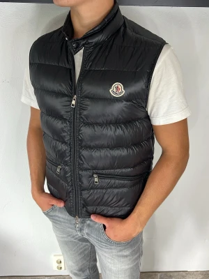 Moncler Gui väst - Snygg svart dunväst från Moncler med hög krage och två dragkedjeförsedda fickor framtill. Västen har ett quiltat mönster och Moncler-logga på bröstet. Perfekt att slänga över en hoodie eller t-shirt för en clean och trendig look.