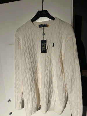 Vit kabelstickad tröja Polo Ralph Lauren - Kabelstickad vit tröja från Polo Ralph Lauren med rund halsringning och klassisk logga broderad på bröstet. Tröjan har lång ärm och ribbade muddar. Perfekt för dig som gillar en stilren och tidlös look.är i storlek xl men passar L/M syd i halsen men det syns knappt