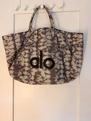 Tie-dye shoppingväska  - Stor shoppingväska i tyg med gråbrunt och beige tie-dye mönster. Väskan har två handtag och stor svart Alo-logga på framsidan. Perfekt för att bära med sig allt man behöver. Riktigt trendig och rymlig design.