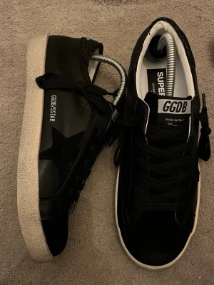 Svarta GGDB sneakers med stjärndetalj - Svarta sneakers från Golden Goose med vit sula och svart stjärndetalj på sidan. Skorna har svarta skosnören, GGDB-logga på plösen och texten GGDB/STAR på sidan. Materialet är en mix av läder och mocka med en klassisk låg siluett.