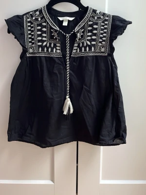 Svart broderad blus från H&M - Svart blus från H&M med vita broderade detaljer och snörning i halsen. Blusen har korta ärmar med volang och ett bohemiskt mönster upptill. 