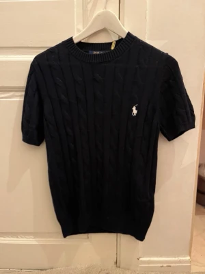 Marinblå kabelstickad topp Polo Ralph Lauren - Kabelstickad marinblå topp från Polo Ralph Lauren med korta ärmar och rund halsringning. Klassisk vit logga broderad på bröstet. Perfekt till jeans eller kjol för en clean och stilren look.Priset går att diskutera vid snabb affär, finn de frågor så är de bara o skriv 😊