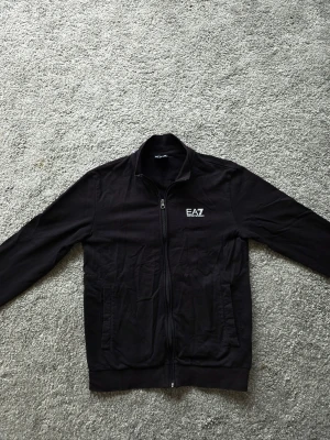 Svart ziptröja från Emporio Armani - Snygg svart ziptröja från Emporio Armani EA7 med hög krage och dragkedja framtill. Tröjan har två fickor på sidorna och en stilren logga på bröstet. Perfekt för en avslappnad och trendig look.