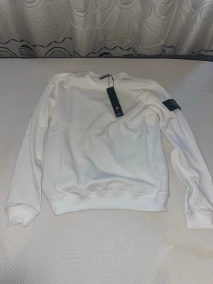 Vit sweatshirt från Stone Island - Vit sweatshirt från Stone Island med rund halsringning och klassisk logotyp-patch på vänster ärm. Tröjan har ribbade muddar vid ärmslut och nederkant. Perfekt för en stilren och avslappnad look.