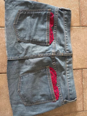 Kort jeanskjol med röda spetsdetaljer - Säljer en kort jeanskjol i blå denim med stora bakfickor och röda spetsdetaljer som sticker fram ur fickorna. Kjolen har en klassisk femficksmodell och rå kant nertill för en avslappnad look.