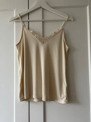Beige linnetopp med spets från H&M - Säljer en beige linnetopp från H&M med tunna axelband och v-ringning dekorerad med spets. Toppen har en enkel och stilren design som passar perfekt till många olika looks. Materialet känns mjukt och lätt mot huden.