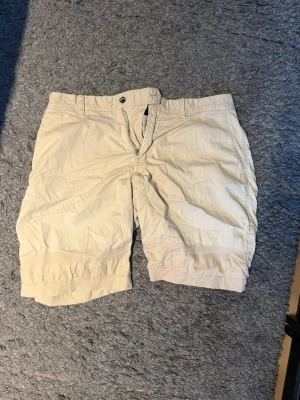 Beige chinos shorts från J.Lindeberg - Snygga beige chinos shorts från J.Lindeberg med klassisk passform och dragkedja framtill. Perfekta för varma dagar och enkla att matcha med olika stilar. Gjorda i mjuk bomull för en skön känsla.
