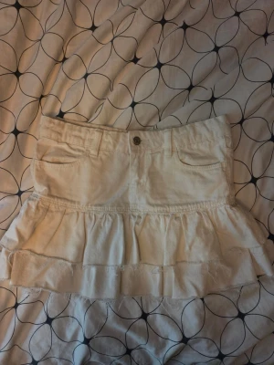 Vit volangkjol med fickor - Kort vit kjol i bomull med volangdetaljer och råa kanter. Kjolen har två framfickor och knappstängning framtill. Perfekt för dig som gillar en avslappnad och trendig stil.