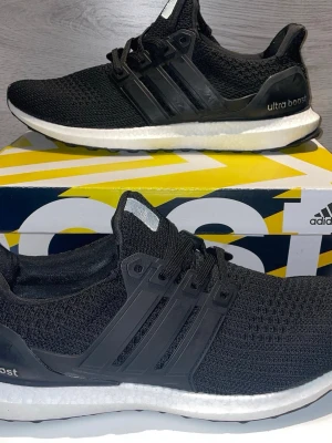 Svarta Adidas Ultra Boost sneakers - Snygga svarta Adidas Ultra Boost sneakers med stickad ovandel i mesh, klassiska tre ränder på sidan och vit Boost-sula för extra komfort. Skorna har en sportig look och passar perfekt till streetwear. Svart plastdetalj runt hälen och snörning framtill.