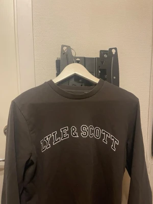 Svart sweatshirt från Lyle & Scott - Cool svart sweatshirt från Lyle & Scott med vit text på bröstet. Tröjan har rund halsringning och långa ärmar. Perfekt till ett avslappnat streetwear-look och enkel att matcha med jeans eller joggers. Stor S