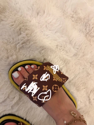 Bruna slides med monogram och gul kant - Snygga slides med bred brun rem dekorerad med mönster i beige. Sulan är svart med en gul kant runt om. Perfekta för en trendig och avslappnad look. Storlek 39, inte använda. ❤️