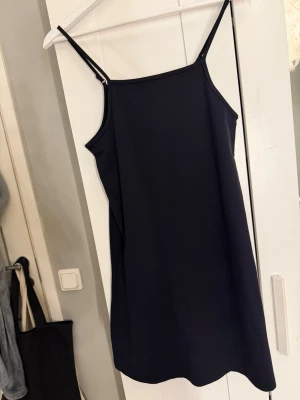 Svart kort klänning med tunna band - En enkel och stilren svart kort klänning med tunna axelband och rak passform. Storlek 158/164 vilket motsvarar Xs. Klänningen har minimalistisk design och är perfekt för dig som gillar en clean look. Materialet ser ut att vara mjukt och följsamt.