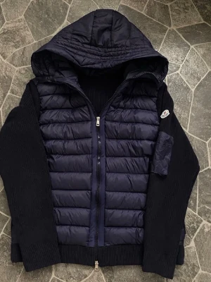 Mörkblå pufferjacka från Moncler - Snygg mörkblå pufferjacka från Moncler med stickade ärmar och huva. Jackan har quiltad front i glansigt material, dragkedja framtill och Moncler-logga på ärmen. Perfekt för kyliga dagar när du vill ha både stil och värme.