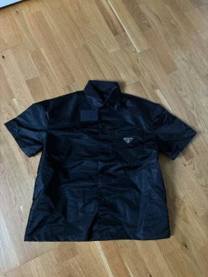 Prada button up T-shirt skjorta  - Snygg svart skjorta från Prada i glansigt material med korta ärmar och klassisk krage. Skjortan har knappar framtill och en bröstficka med Pradas ikoniska metallogo. Perfekt för dig som gillar exklusiv och stilren design.