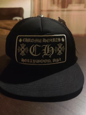 Svart Chrome Hearts keps - Snygg svart keps från Chrome Hearts med broderad patch framtill där det står 'CH' och 'Hollywood, USA'. Kepsen har böjd skärm och mesh baktill för extra ventilation. Justerbar storlek med snapback-spänne. Pris går såklart och förhandlas under affär