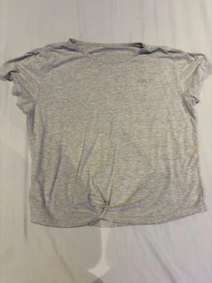 Ljusgrå t-shirt - En basic ljusgrå t-shirt med korta ärmar och rund halsringning. T-shirten har en enkel design och är tillverkad i mjukt material, perfekt för en avslappnad stil. Har knytning vid midja som ger en fin touch.