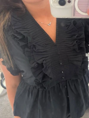 Svart blus med volanger och v-ringning - Supersnygg svart blus med djup v-ringning och stora volanger framtill. Blusen har korta ärmar och dekorativa knappar längs bröstet. Materialet känns lätt och luftigt, perfekt för att lyfta vilken outfit som helst.