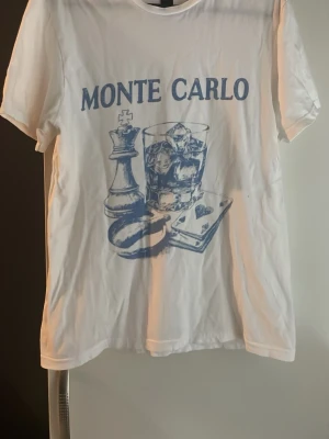 Vit t-shirt med Monte Carlo-tryck - Vit t-shirt med blått tryck som föreställer texten 'Monte Carlo', ett glas med is, schackpjäs och spelkort. Klassisk rund hals och korta ärmar. Perfekt för dig som gillar unika motiv och en avslappnad stil.