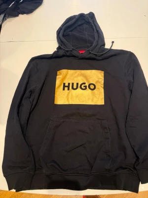 Svart hoodie med guld HUGO-logga - Snygg svart hoodie med stor guldig HUGO-logga på bröstet. Har ett jätte litet hål  