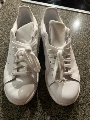 Vita Adidas Stan Smith sneakers, stl 39 1/3 - Klassiska vita Adidas Stan Smith sneakers i lågt snitt med vita skosnören och diskret perforering på sidorna. Tillverkade i slätt läder med präglad logga och porträtt på plösen. Perfekta för en clean och stilren look. Endast använda en gång, som nya! 