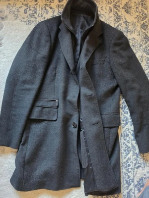 Mörkgrå ullrock från Balmain - Stilren mörkgrå rock från Balmain i en exklusiv mix av ull och kashmir. Klassisk design med krage, knappstängning och flera fickor, inklusive en med dragkedja. Perfekt för dig som vill ha en tidlös och elegant look.