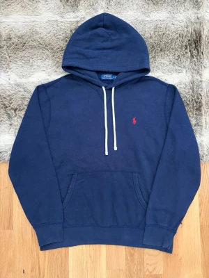 Mörkblå hoodie från Polo Ralph Lauren - Mörkblå hoodie från Polo Ralph Lauren med vit dragsko i huvan och klassisk röd logga broderad på bröstet. Hoodien har känguruficka framtill och ribbade muddar vid ärmar och nederkant. Perfekt för en avslappnad och stilren look.