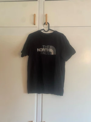 Svart t-shirt från The North Face - Snygg svart t-shirt från The North Face med stor logga i vitt och grått på bröstet och liten logga på ryggen. Klassisk passform och rund halsringning. Perfekt till jeans eller shorts för en avslappnad look.
