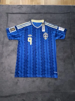 Blå Sverige fotbollströja Adidas - Säljer en blå Sverige fotbollströja från Adidas med gula detaljer och nummer 9 på bröstet. Tröjan har ett vågigt mönster, broderad svensk landslagssköld och tre gula ränder på axlarna. Tillverkad i lätt och ventilerande material.