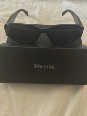 Svarta solglasögon från Prada - Snygga svarta solglasögon från Prada med rektangulära bågar och mörka glas. Klassisk och stilren design med breda skalmar. Kommer med originalfodral med Prada-logga.