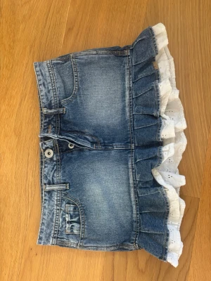 Jeanskjol med volang och spets - Söt kort jeanskjol med volangkant i blå denim och vit spetsdetalj längst ner. Kjolen har klassiska fickor fram och bak samt knapp- och dragkedjestängning. Perfekt för sommaren 