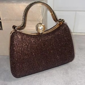 Brun glittrig handväska med pärldetalj (oanvänd ) - Snygg brun glittrig handväska perfekt för fest! Den har en elegant pärldetalj på låset och ett glittrande handtag. Väskan kommer med en avtagbar kedja så du kan bära den som axelväska också. Perfekt accessoar för att lysa upp din outfit! Oanvänd köpt till bröllopsfest