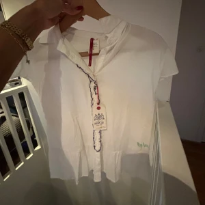 Vit blus från Replay - Säljer en superfin vit blus från Replay. Den har korta ärmar och en söt volangdetalj längs knäppningen. Perfekt för både vardag och fest! Blusen är i nyskick och har en luftig passform.