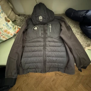 Svart jacka från Sail Antarctic - Säljer en svart jacka från Sailracing Antarctic. Den har en cool design med ett märke på ärmen och dragkedja framtill. Jackan är perfekt för kyligare väder och har en quiltad stil som ger extra värme. Den är i bra skick och redo för nya äventyr! 