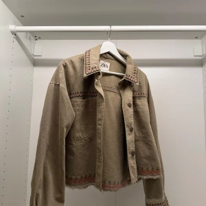Beige jacka från Zara - Säljer en superfin grön jacka från Zara i storlek S. Den har snygga broderade detaljer i rött och grönt längs kragen, ärmarna och nederkanten. Jackan är croppad och har knappar framtill. Perfekt för höst och vår!