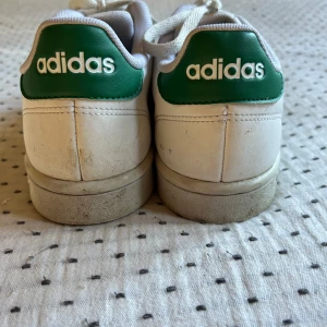 Vita Adidas sneakers med gröna detaljer - Säljer ett par vita Adidas sneakers med gröna detaljer. De är lite smutsiga men fortfarande i bra skick. Perfekta för vardagsbruk med klassisk design och bekväm passform. Skorna har snörning och är gjorda i syntetmaterial. Lite söndriga vid häl sula.