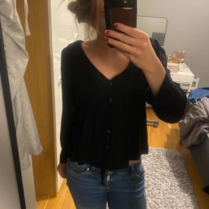 Svart blus med knappar - Säljer en svart blus med knappar framtill. Den har en v-ringad design och långa ärmar, perfekt för både vardag och fest. Blusen  är i ett mjukt material och har en skön passform. Passar bra till jeans eller kjol! Från na-kd i storlek 34💕💕🤗