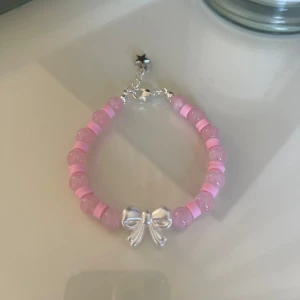 Sött rosa armband med rosett - Det här är ett supergulligt armband med rosa pärlor och en söt liten rosett i silverfärg. Perfekt för att ge en extra touch till din outfit! Armbandet har också en liten stjärna vid låset som ger det en unik detalj.