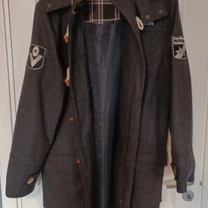 Mörkgrå duffeljacka med träknappar - Unik snygg knappt använd Duffeljacka från Australien. Märkena är emblem för ett Australiensiskt Fotbollslag. Jag har låtit sy dit innefickor