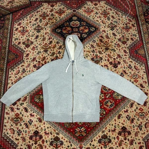 Polo Ralph Lauren zip hoodie - Grå Polo Ralph Lauren zip hoodie grön häst. Hoodien har två fläckar, man tänker inte på dom alls. Ena fläcken (sista bilden längst ner) e på baksidan av tröjan längst ner på sömmen. Andra är högt på ryggen sminkfläck som knappt syns. Annars bra skick