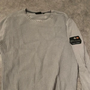 Grå stickad tröja från Sail Racing - Säljer en stilren grå stickad tröja från Sail Racing. Tröjan har en rund halsringning och en diskret logga på ärmen. Perfekt för kyligare dagar. 