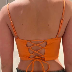 Orange korsettopp med snörning - Trendig orange korsettopp med smala axelband och snygg snörning i ryggen. Toppen är croppad och har en figurnära passform som framhäver silhuetten.